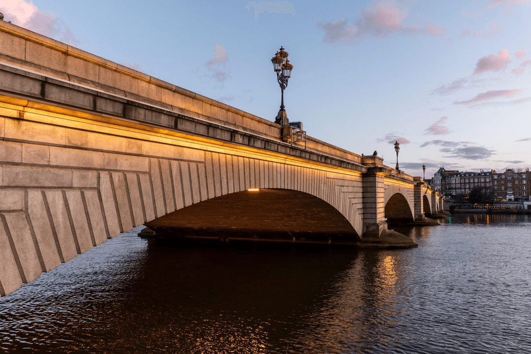 Putney Bridge lit at dusk with golden lamp posts reflected in the darkening Thames below